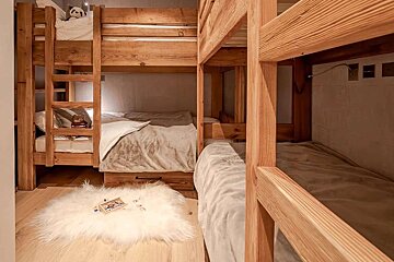 A cozy room features several wooden bunk beds with soft bedding and plush toys. A fluffy white rug lies on the light wooden floor.