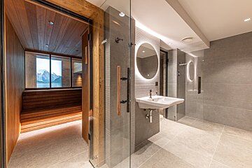 A modern spa area features a wooden sauna with mountain views through a window, adjacent to a sleek bathroom with an illuminated mirror and sink.
