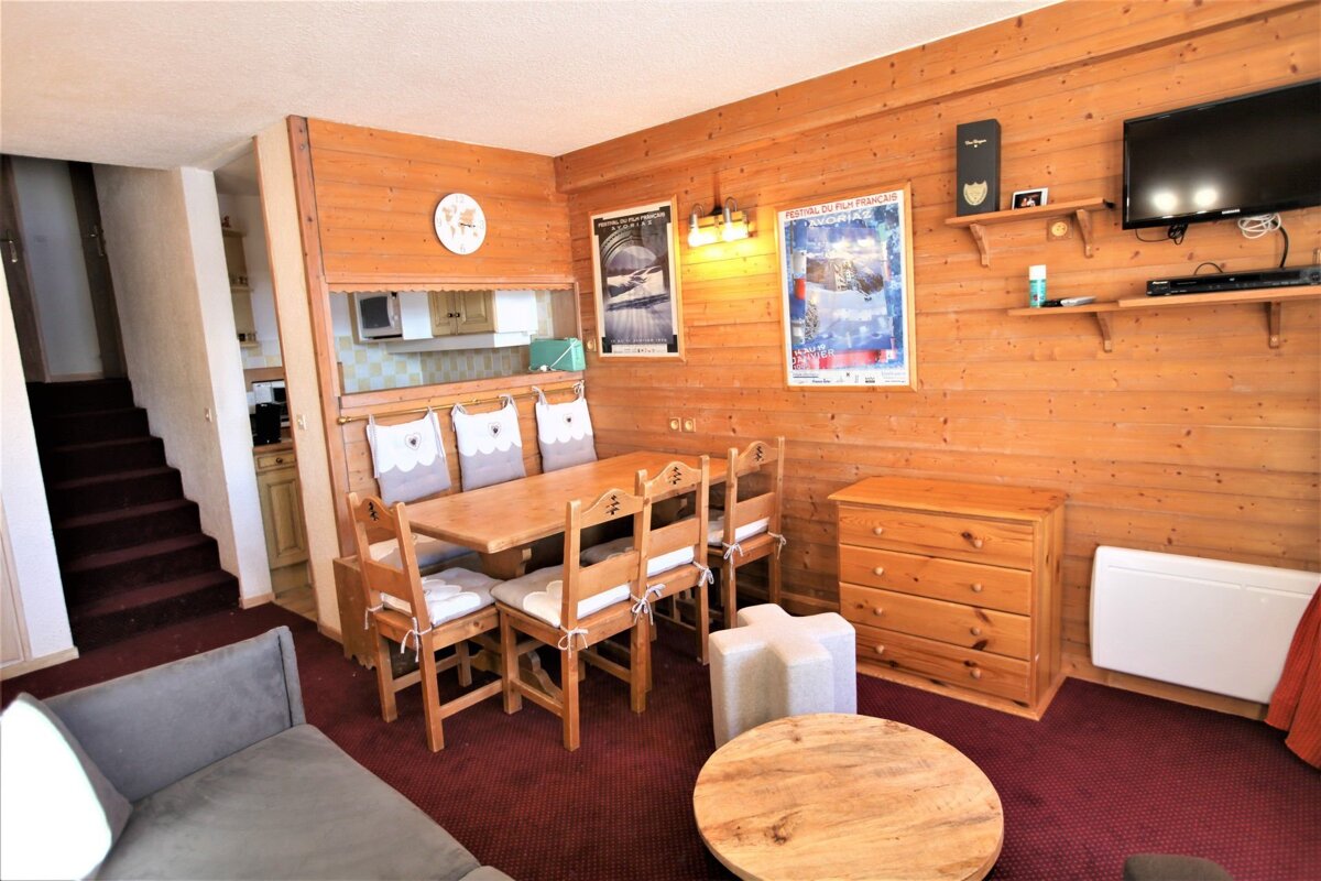 A cozy, wood-paneled living/dining room with a wooden dining set, red carpet, and a view into a kitchenette. Stairs are visible on the left.