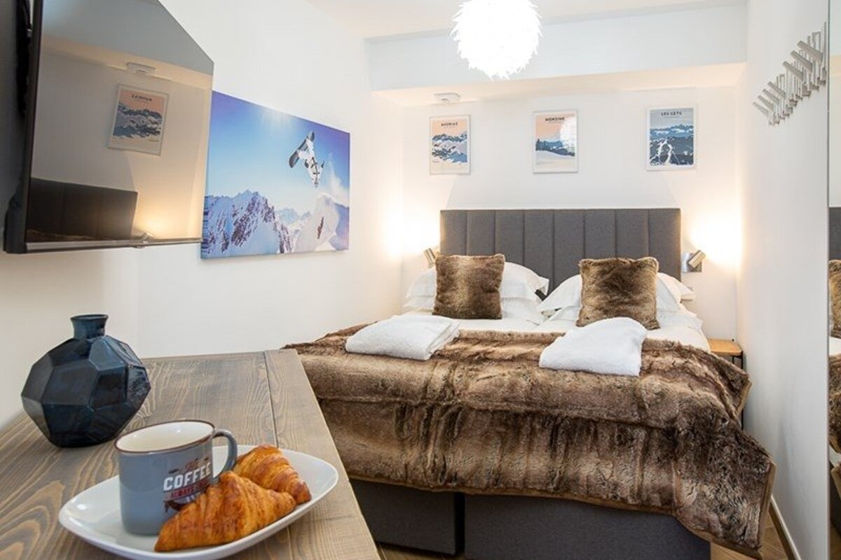 A cozy ski-lodge bedroom featuring a fur blanket, mountain art, and a breakfast tray with croissants and coffee in the foreground.
