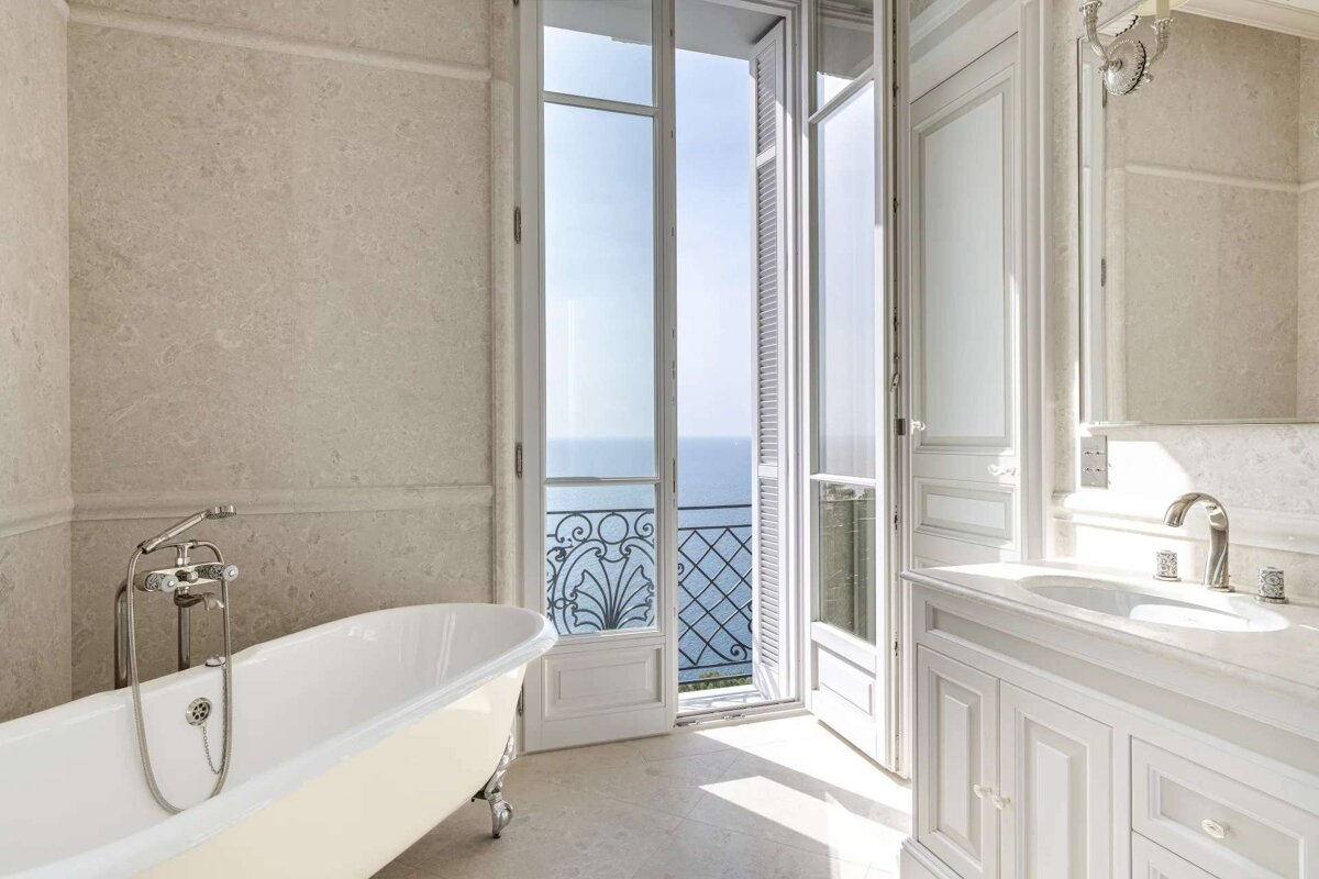An elegant, sunlit bathroom featuring a clawfoot tub, marble vanity, and French doors opening to a stunning balcony with an ocean view.