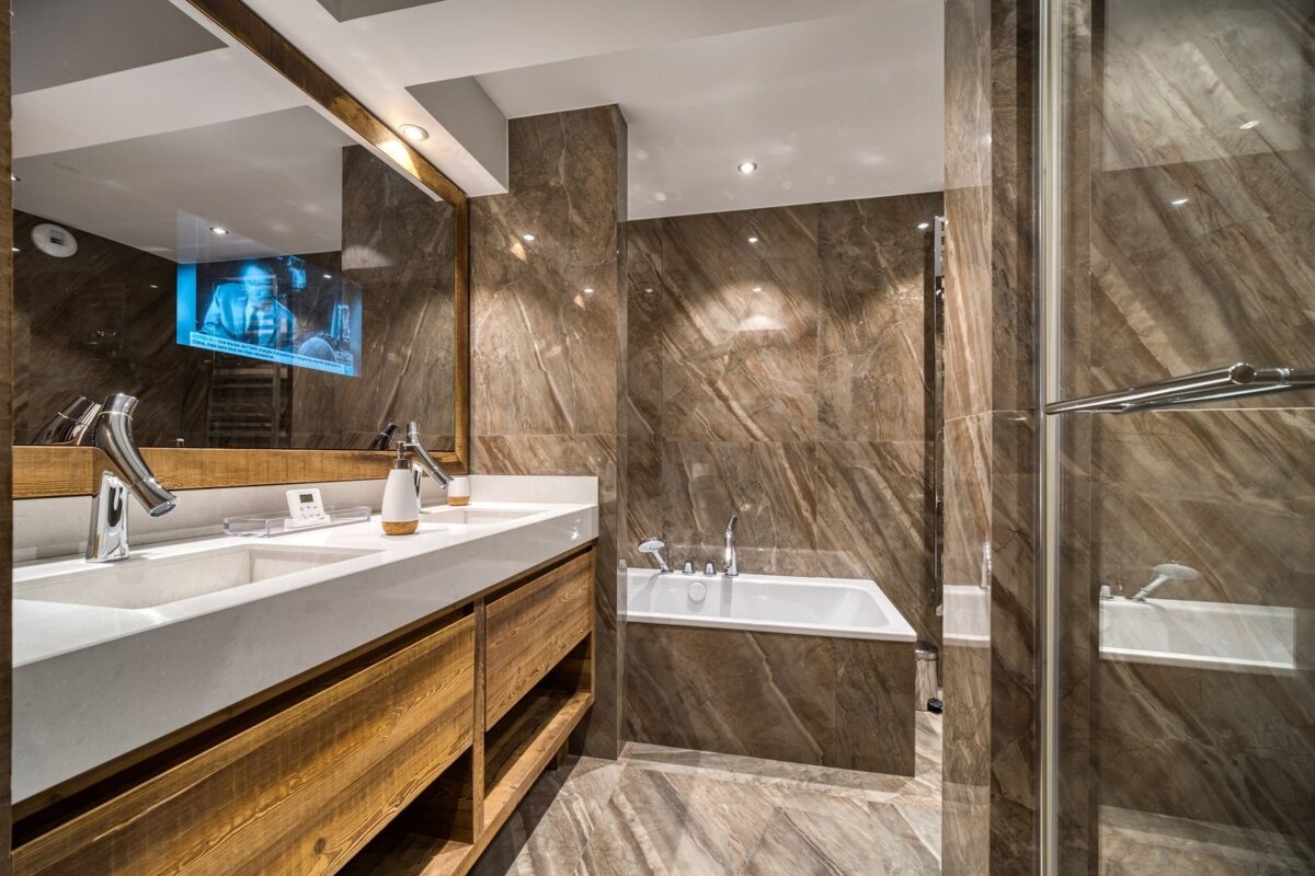 Luxurious marble bathroom with double wood vanity, modern fixtures, a bathtub, glass shower, and a TV integrated into the mirror.