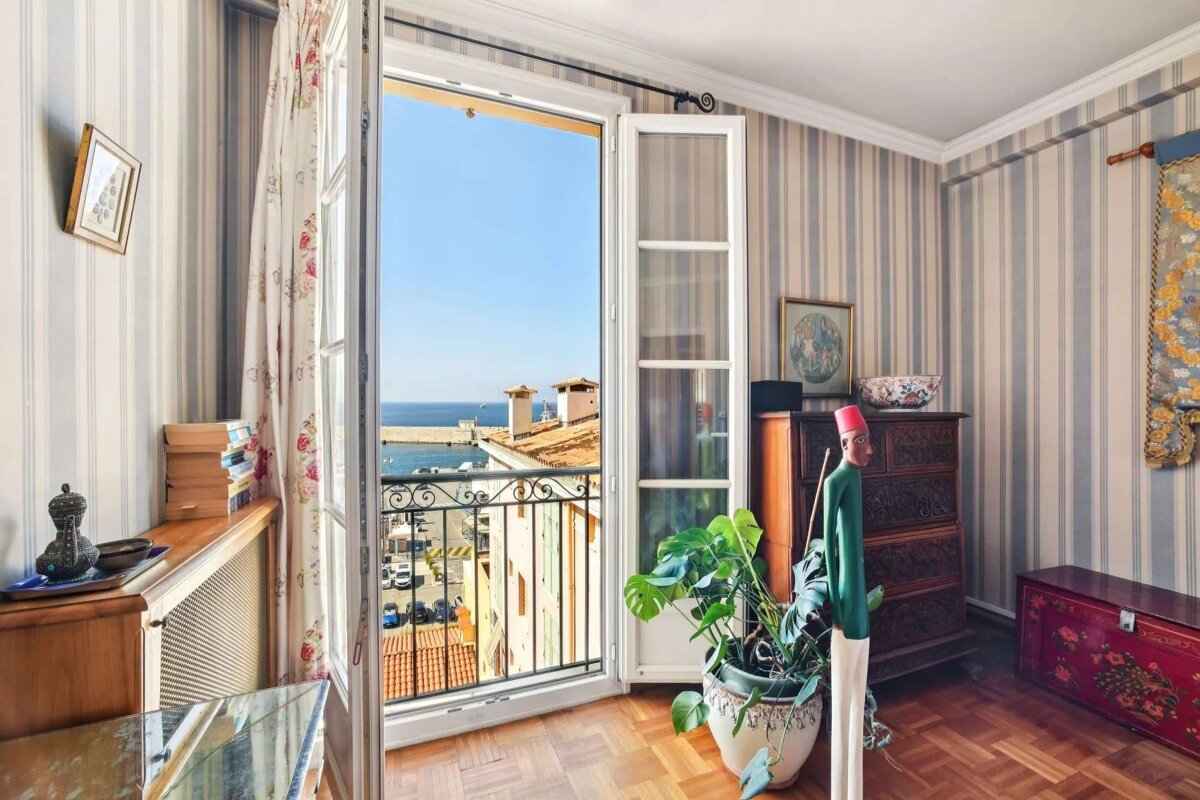 A charming room with striped walls and wooden floors, offering an open balcony view of the sunny blue sea and a bustling harbor below.