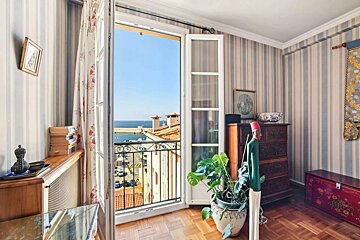 A charming room with striped walls and wooden floors, offering an open balcony view of the sunny blue sea and a bustling harbor below.