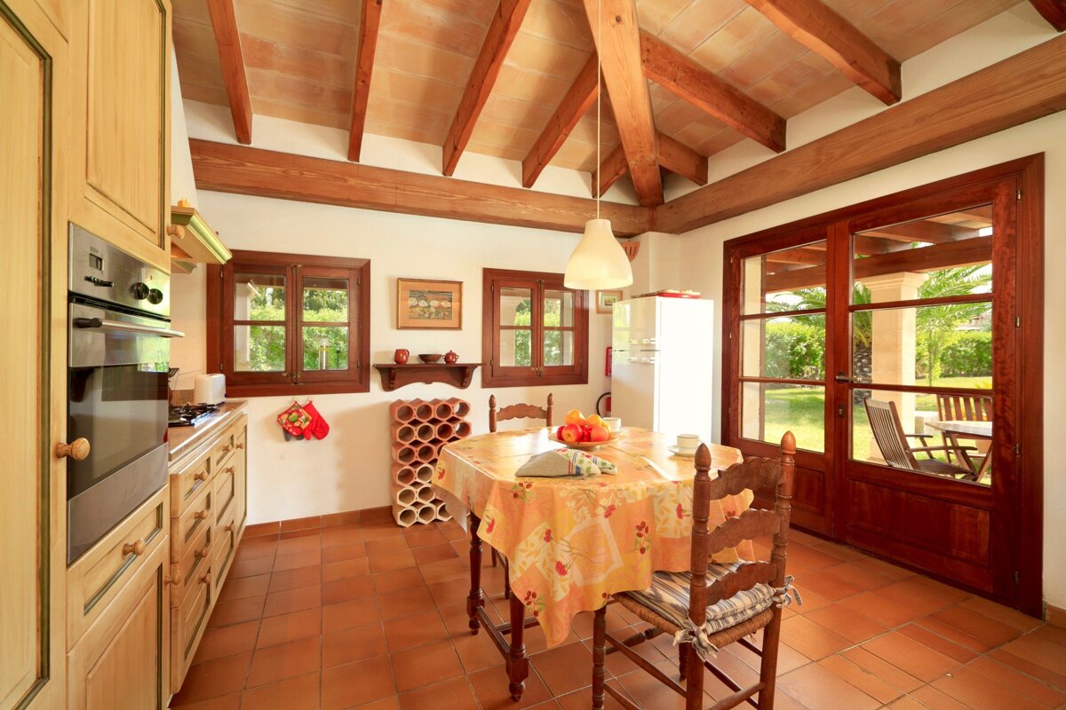 Cozy, rustic kitchen featuring exposed wooden beams, terracotta floor, a dining area, and glass doors opening to a bright garden.