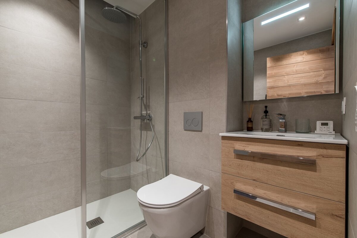 Modern grey-tiled bathroom with a glass walk-in shower, toilet, and a light wood vanity featuring a sink and illuminated mirror.