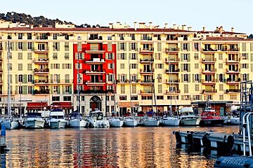 A vibrant harbor scene featuring boats docked before colorful buildings with yellow and a prominent red section, all reflected in the calm water.