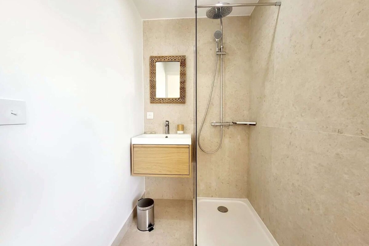 A modern bathroom features a glass-enclosed shower, a floating wooden vanity with a sink, and a decorative mirror, all against light-toned walls.