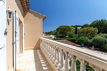 Sunny balcony with white balustrade and shutters, overlooking a vibrant green landscape under a bright blue sky.