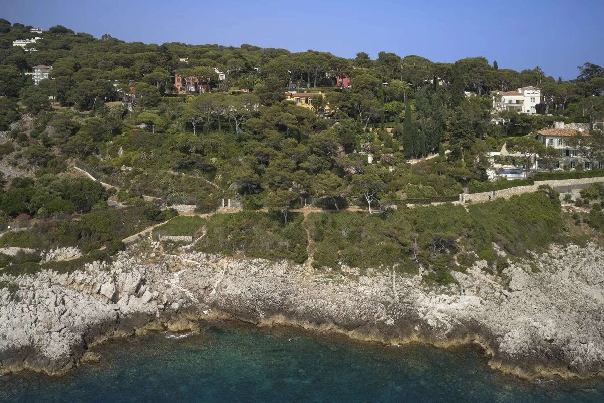 Rocky coastline with clear blue water, a densely forested hill dotted with houses, all under a bright sky.