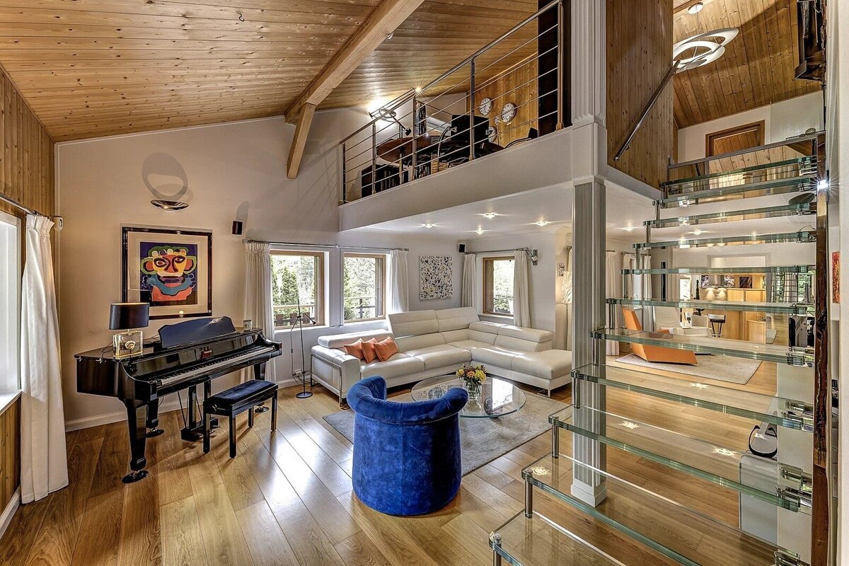Spacious, modern living room with a grand piano, white sectional sofa, blue chair, and a striking glass staircase leading to a mezzanine.