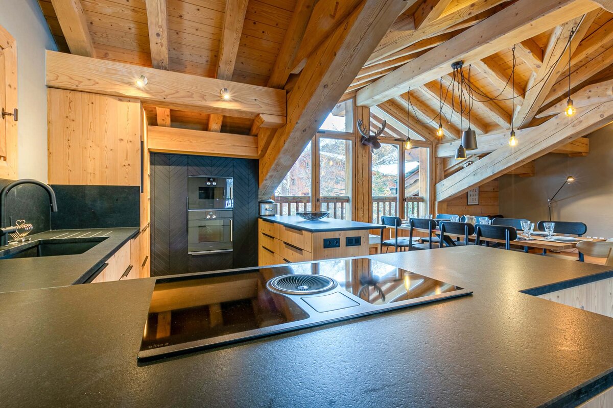 A stylish, open-plan kitchen and dining area in a modern wooden chalet, featuring exposed beams, a sleek cooktop, and large windows.