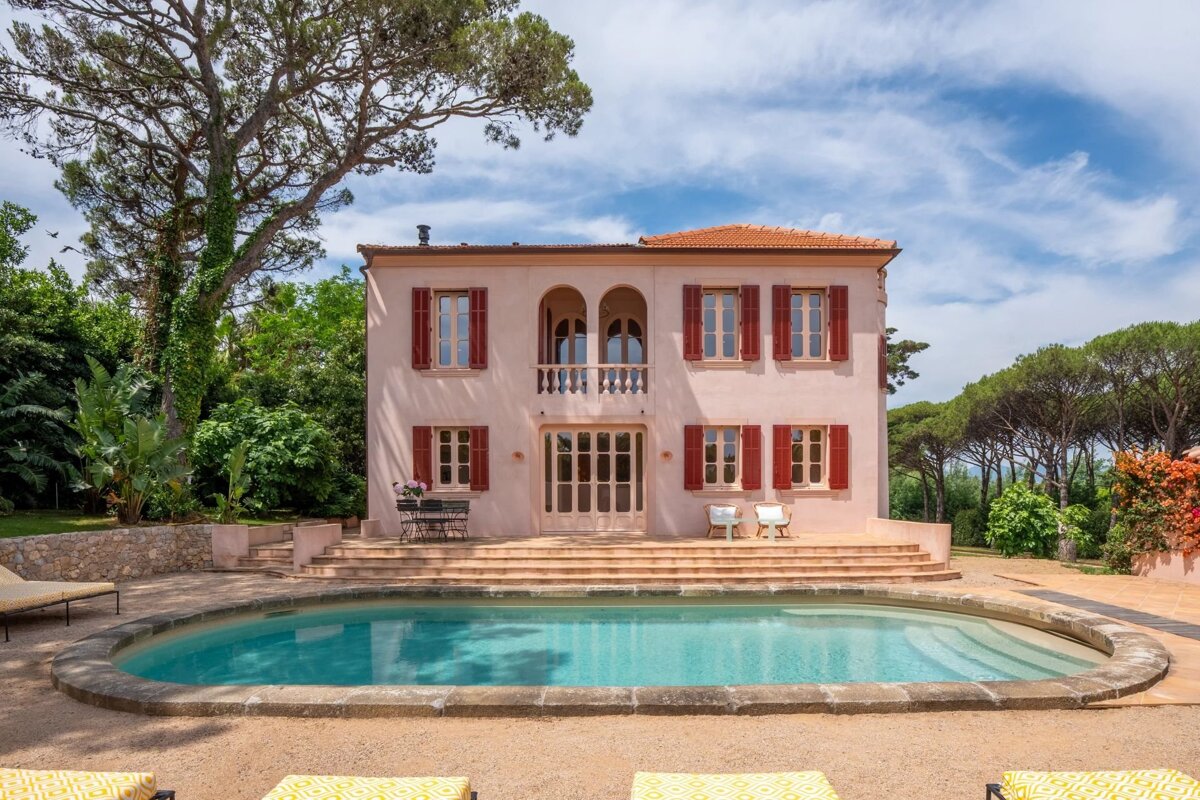 An elegant pink Mediterranean villa with red shutters, fronted by a curved swimming pool and surrounded by trees.