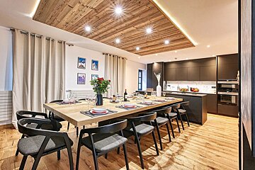Modern dining area and open kitchen with a long wooden table, dark chairs, and a planked wooden ceiling with recessed lighting.
