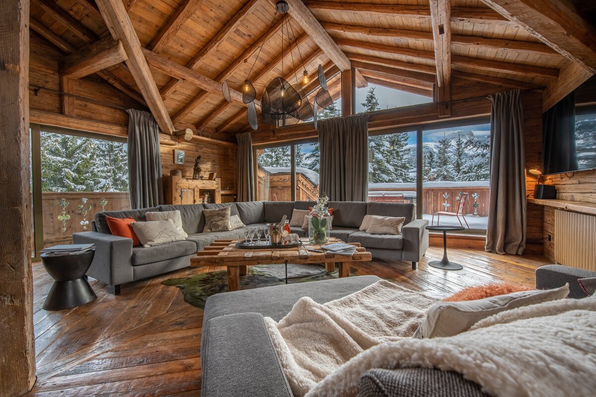 Cozy, rustic wooden chalet living room with large windows overlooking a snowy mountain landscape, featuring comfortable gray sectional sofas.