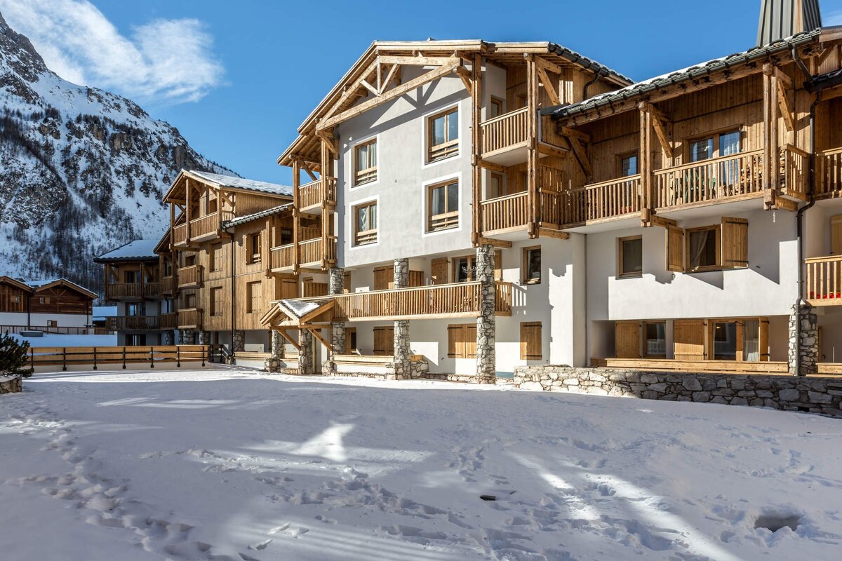 Modern chalet-style buildings with wood balconies, surrounded by snow, under a sunny sky with a mountain backdrop.