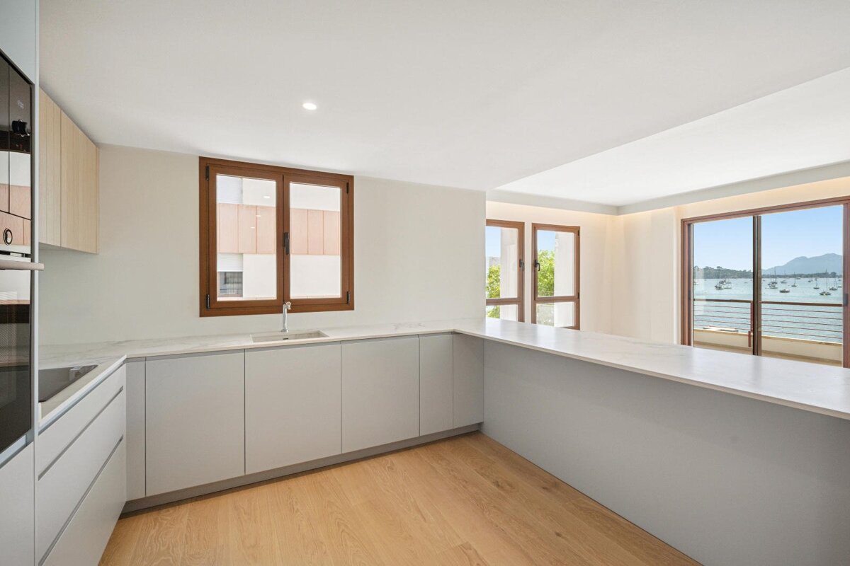 Modern kitchen with light cabinetry, wooden floors, and large windows offering a beautiful sea view with boats and mountains. Bright and open.