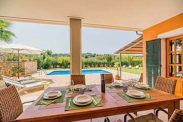 A covered outdoor dining area set for a meal, overlooking a sparkling blue swimming pool and lush green landscape under a bright sky.