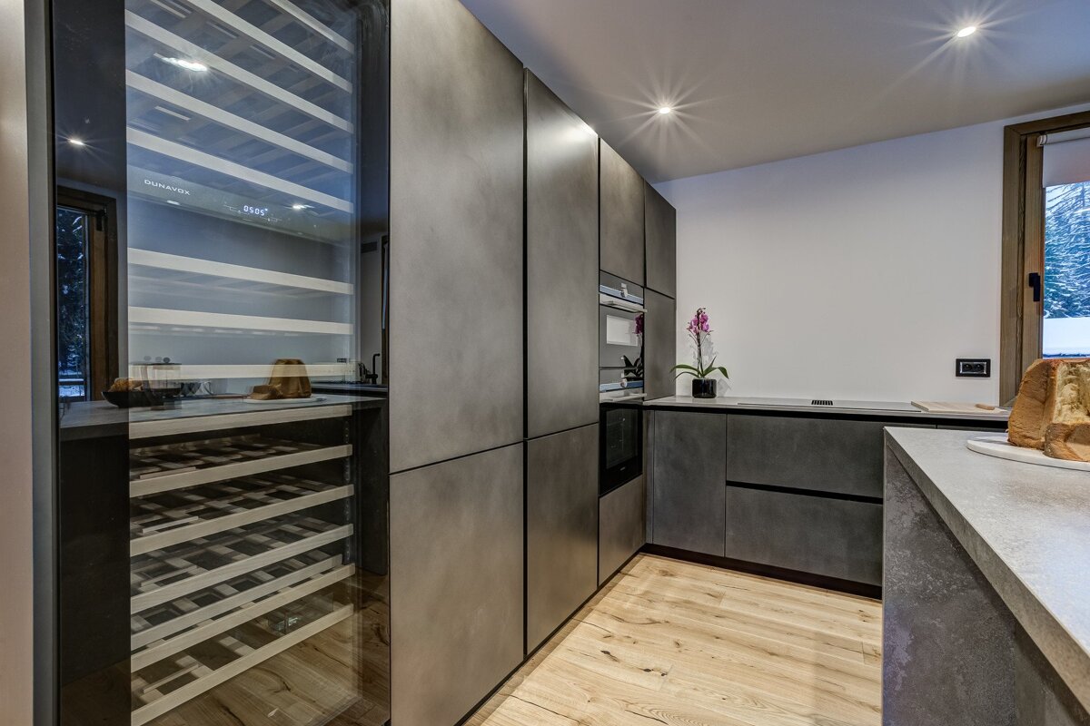 A modern kitchen features dark grey, minimalist cabinetry, light wood flooring, a large wine cooler, and built-in appliances. A snowy outdoor view is seen through a window.
