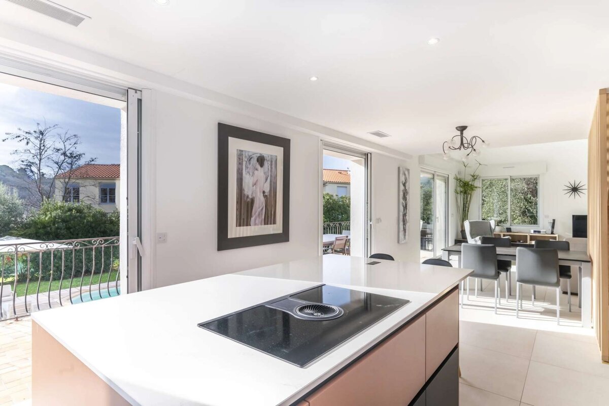 Modern kitchen with a large island stovetop and white counters, connected to a dining area, offering views of an outdoor pool and house.