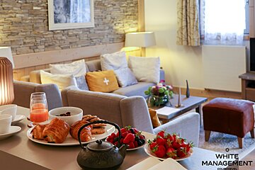 A cozy living room features a table laden with a delightful breakfast spread: croissants, fresh berries, juice, and tea, set against a stone wall and sofas.