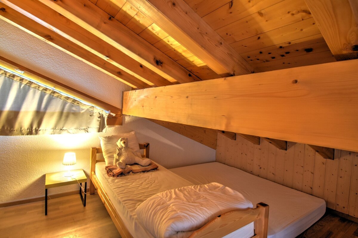 Cozy attic bedroom with wooden beams, two beds (one with a teddy bear), and a bedside lamp, creating a warm ambiance.
