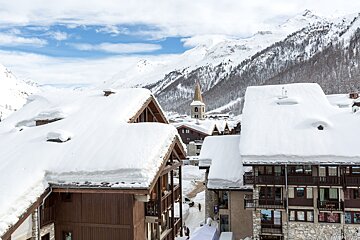 Snow-covered wooden chalets in an alpine village, with a church steeple and majestic snowy mountains in the background.