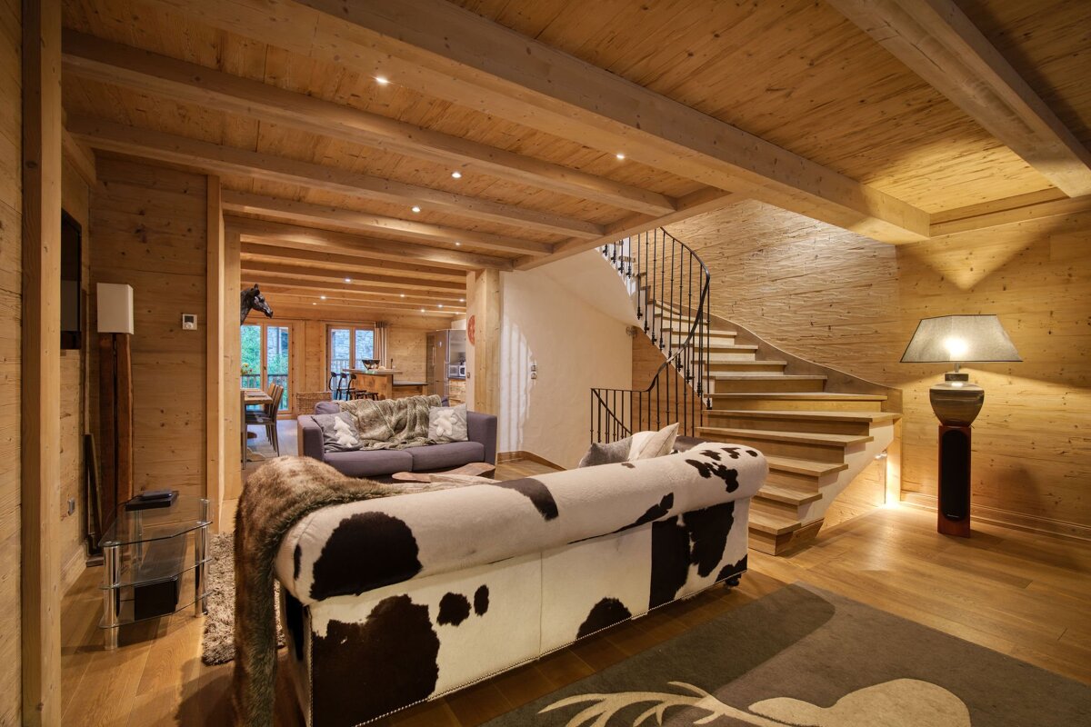 Cozy wooden chalet interior featuring a cowhide sofa, exposed ceiling beams, a winding staircase, and warm lighting.