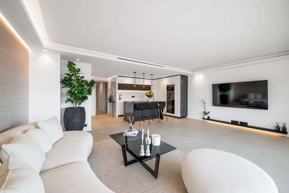 A modern, open-plan living room with a white curved sofa, glass coffee table, and large TV, seamlessly connected to a kitchen with a dark island and bar stools.