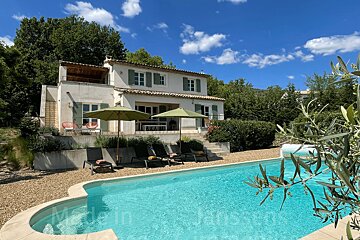 A sunny white villa with green shutters overlooks a sparkling blue swimming pool, flanked by lounge chairs and lush greenery under a bright sky.