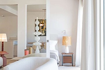 A bright, modern interior with white walls, showing a bedroom with a woven lamp and a glimpse of a living/dining area with abstract art and a sculptural lamp.