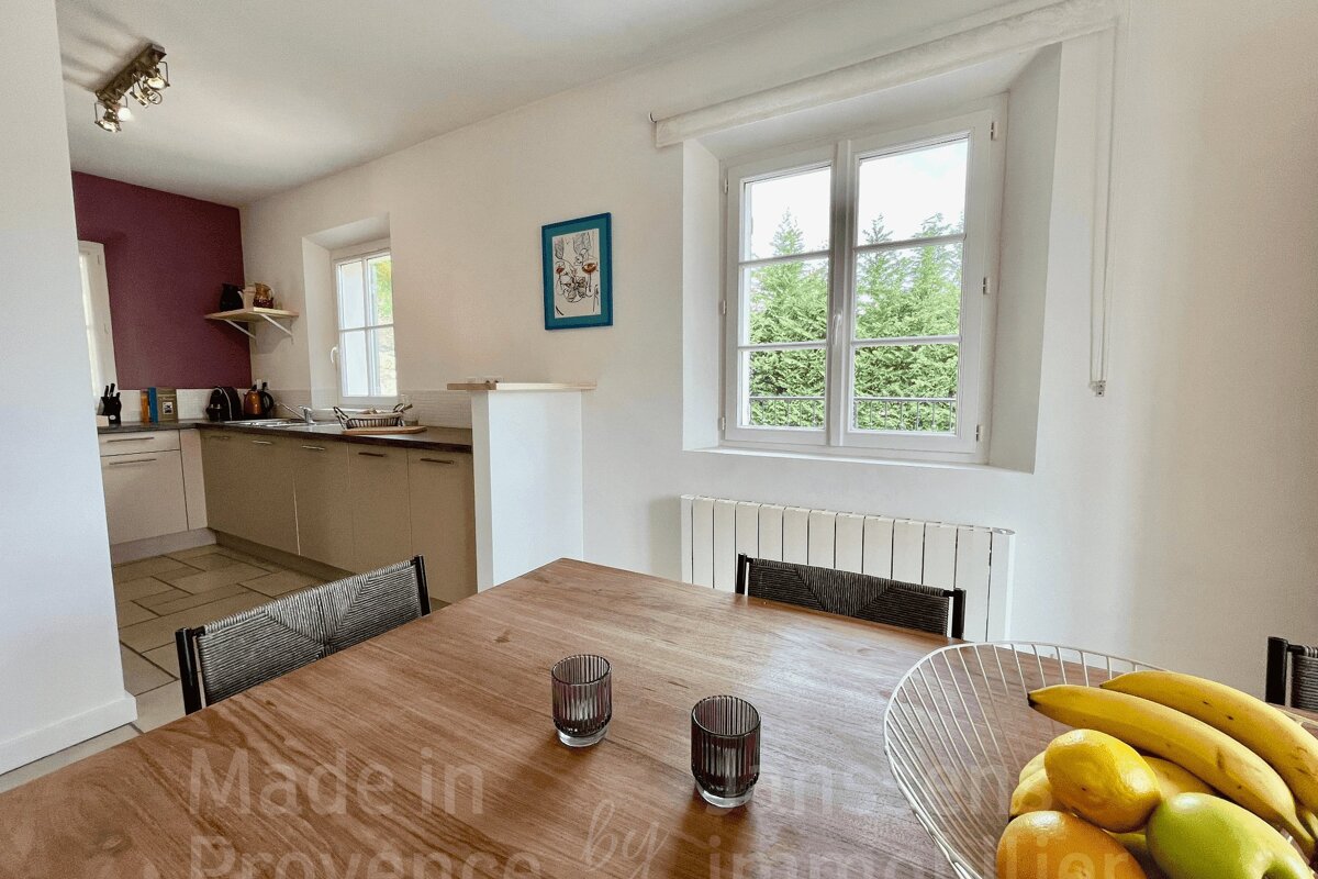 A bright, modern kitchen and dining area with a wooden table, fruit, and window views of green trees. White walls with a purple accent.