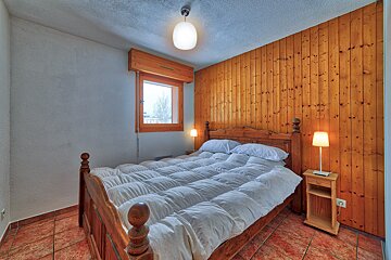 A cozy bedroom with a wooden bed, white duvet, and a striking wood-paneled wall. Features a window, tiled floor, and warm lighting.
