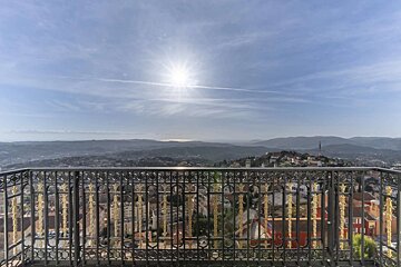 Panoramic view from an ornate balcony, overlooking a sprawling city, distant mountains, and the sea under a bright, sunny sky.