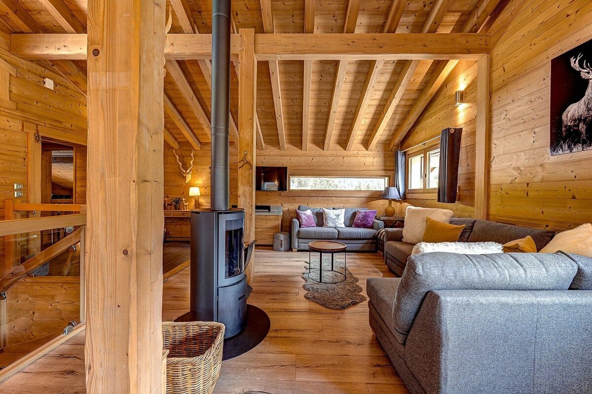 A cozy, modern wood-paneled chalet living room with grey sofas, a freestanding fireplace, exposed beams, and nature-inspired decor.