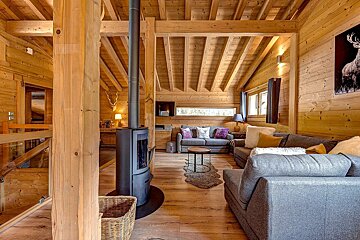A warm, cozy wooden chalet living room features a modern fireplace, grey sofas with colorful cushions, and exposed wooden beams on the ceiling.