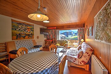 A cozy, wood-paneled chalet living-dining room with checkered table, sofas (one with ski pattern), TV, and balcony view of a mountain village.