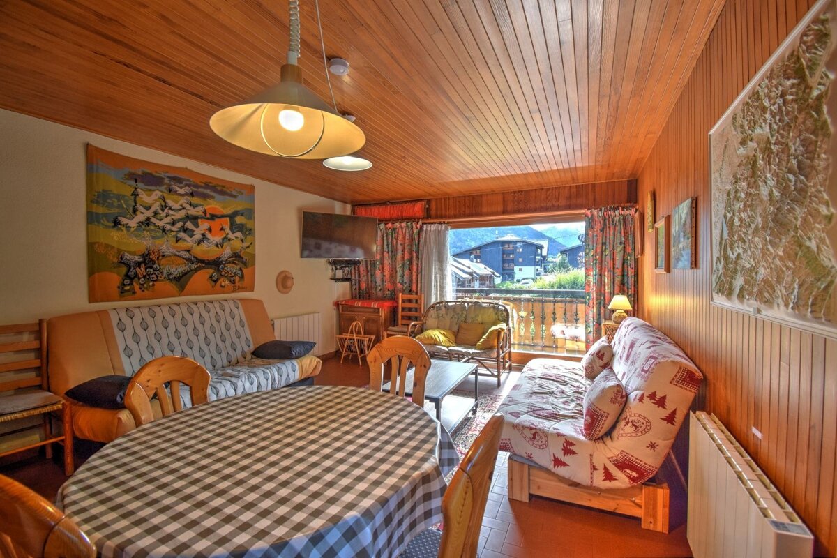 Cozy wood-paneled alpine interior with a checkered dining table, sofas, and a balcony overlooking scenic mountain buildings.