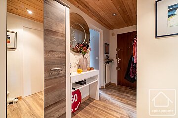 An inviting entryway with wooden ceiling and floor, white walls, a round mirror above a white console with flowers, and a dark wood door.