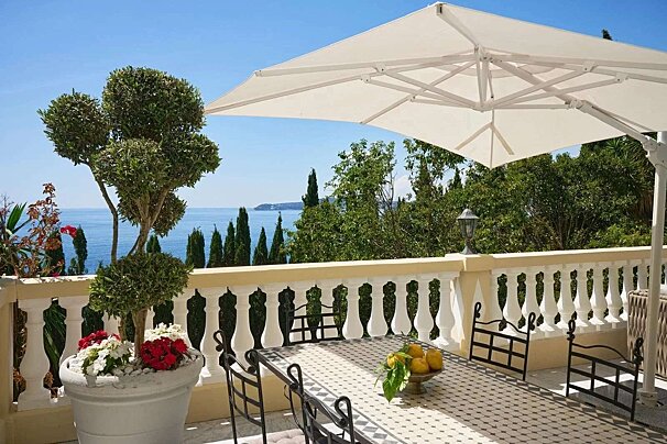 A sunny balcony boasts a dining table under a white umbrella, offering stunning sea views, lush trees, and potted flowers.