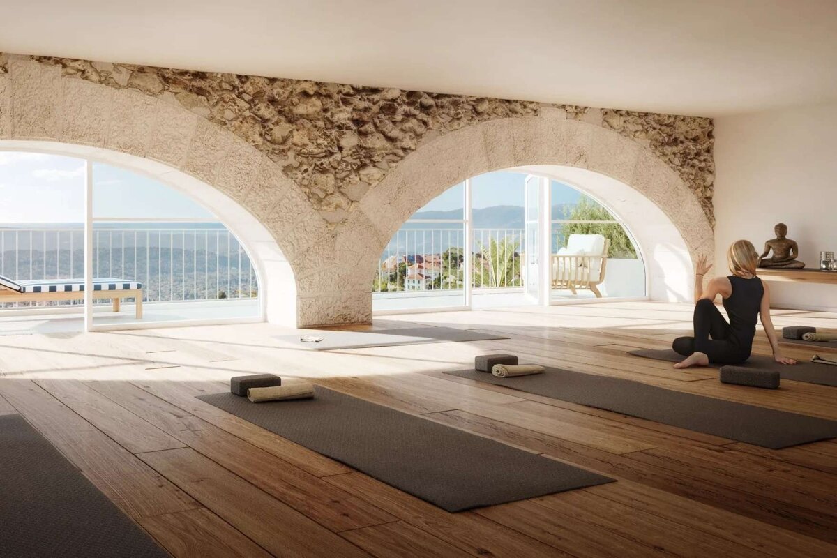 A serene yoga studio with a woman in a pose, featuring rustic stone arches, wooden floors, and expansive windows offering a beautiful sea and mountain view.