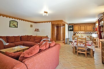 Cozy chalet interior featuring a large reddish sectional sofa, tiled floors, and a wooden dining table with patterned tablecloth.
