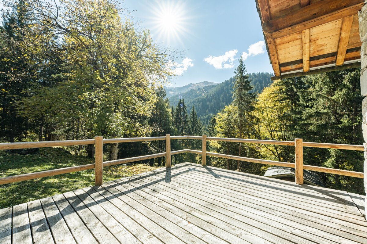 A sunny wooden deck with a fence offers a picturesque view of a vibrant green forest and distant mountains under a bright blue sky.