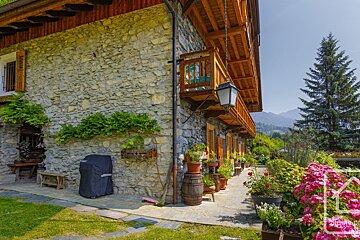 Picturesque stone alpine house with wooden balconies and a vibrant flower garden. Distant mountains rise under a clear blue sky.