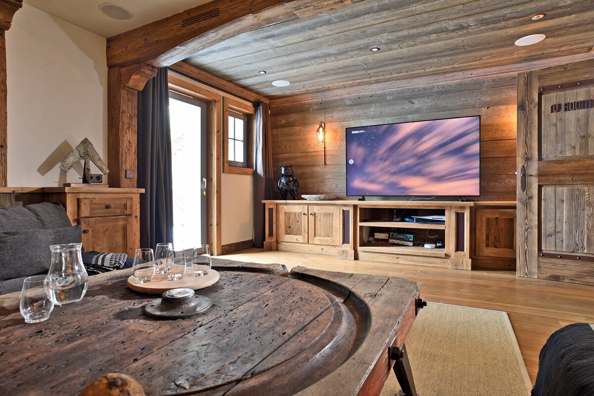 Rustic, wood-filled living room with a modern TV showing a night sky. Features exposed beams, wood-paneled walls/ceiling, and a large antique coffee table.