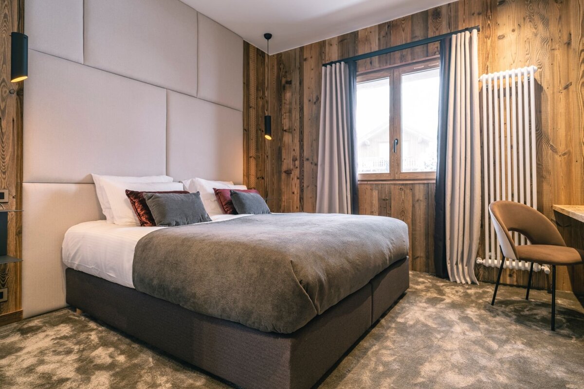 A modern rustic bedroom features a large bed with a padded headboard against wooden wall panels, a window, and a small desk with a chair.