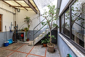 An aged, enclosed courtyard features concrete stairs, potted plants, and tools. A large window reflects the space, while a translucent canopy provides shade overhead.