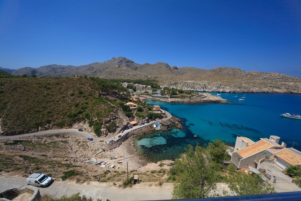 Panoramic view of a stunning coastal bay with turquoise water, mountains, a town, and a winding road. Several boats are in the clear sea.