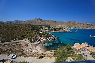 Panoramic view of a stunning coastal bay with turquoise water, mountains, a town, and a winding road. Several boats are in the clear sea.