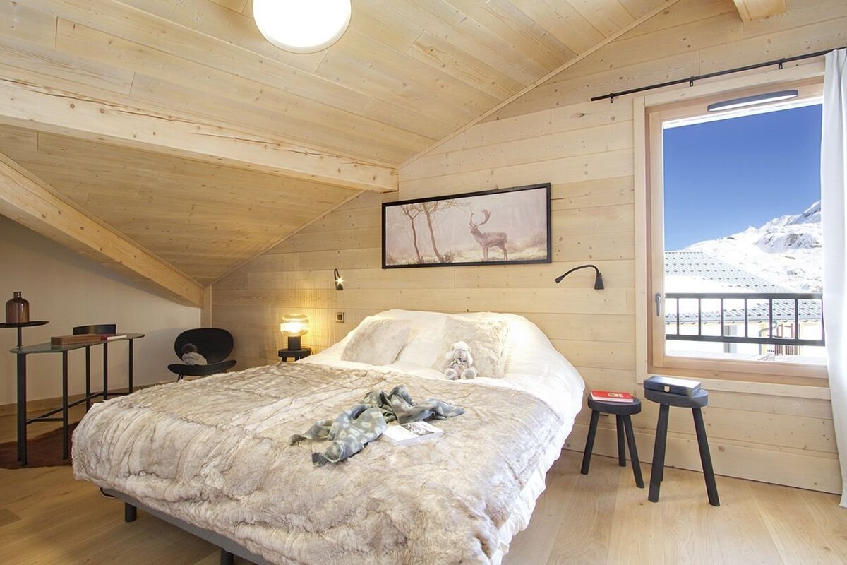 A bright, wood-paneled chalet bedroom features a large bed with a fur throw. A window offers a stunning view of snow-capped mountains.